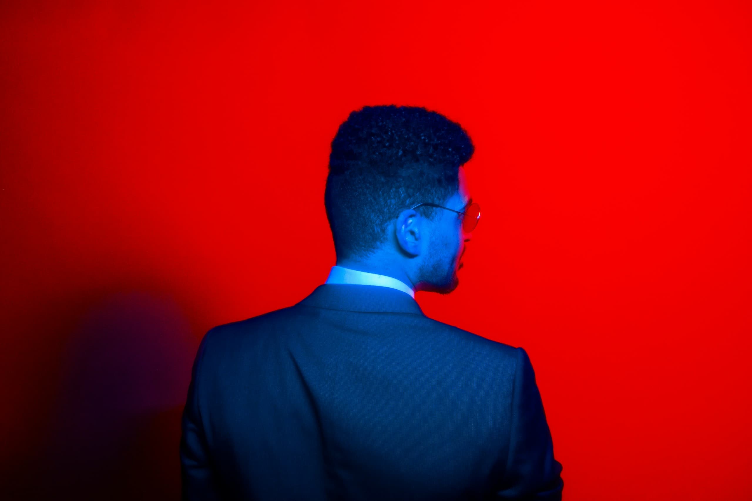 A man in a navy suit, seen from behind, against a saturated red wall
